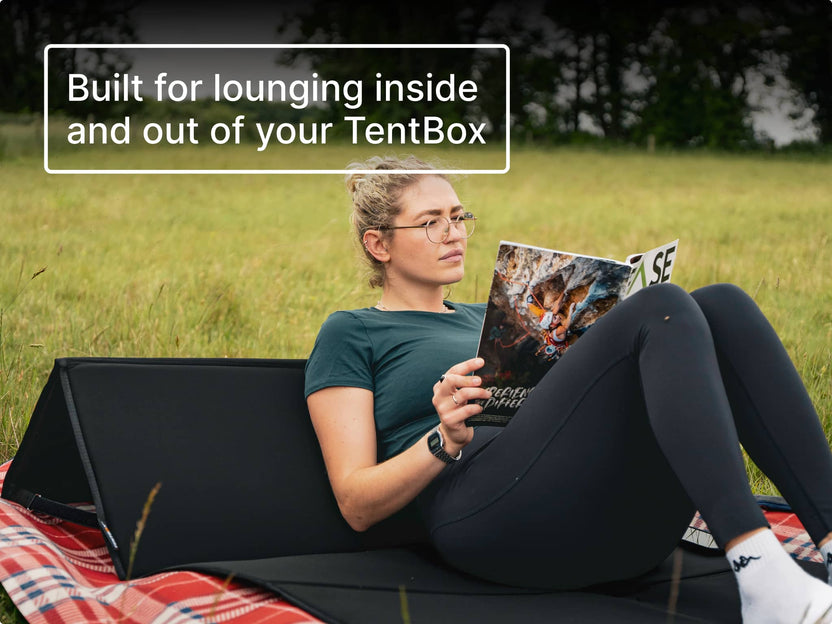 Woman reading a magazine on a blanket in a field, leaning against the TentBox BackRest with text 'Built for lounging inside and out of your TentBox'.