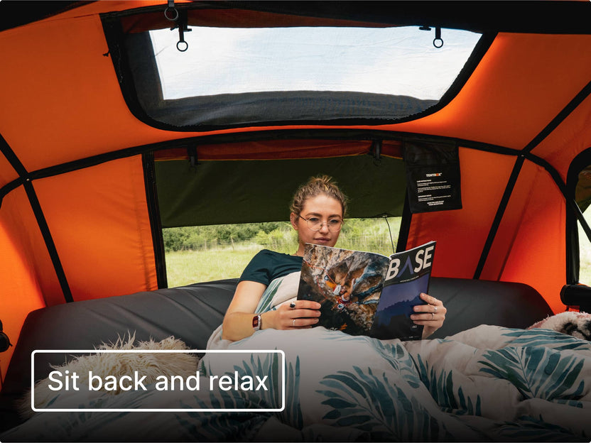 Person reading a magazine inside an orange TentBox Lite XL with a scenic view outside. Text reads 'Sit back and relax'.