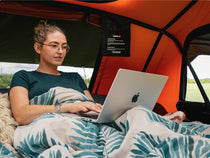 Woman leans against the BackRest inside an orange TentBox, sitting up under a duvet and using her laptop.