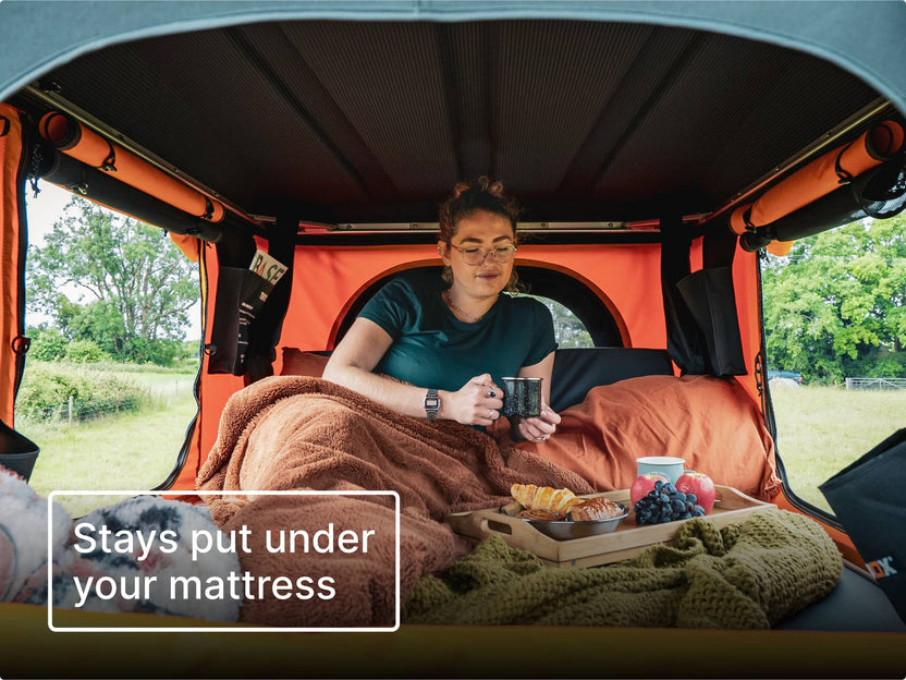 Person sitting inside an orange TentBox Classic under blankets, holding a cup with a tray of breakfast and natural woodland views outside the windows. Text reads 'stays put under your mattress'.